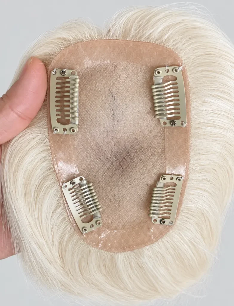 A hand holding a pale blonde hair topper to show the mesh base, smooth edging and four light gold clips.