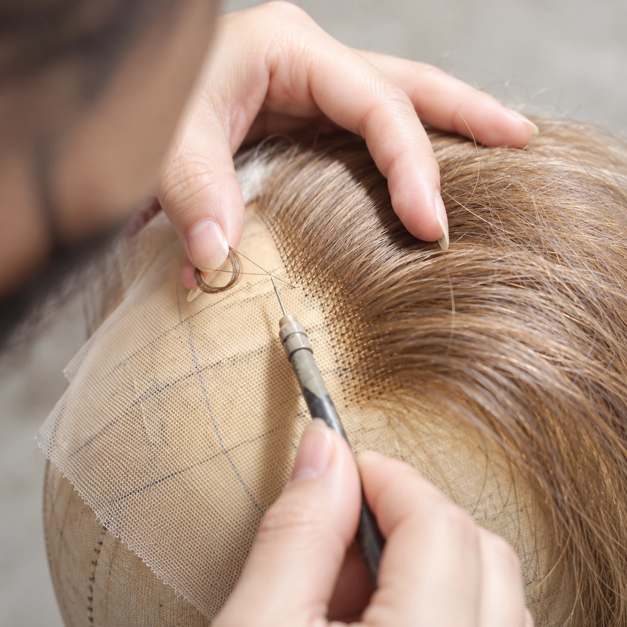 Hands using a fine ventilating hook to knot light brown hair into lace on a fabric-covered support.