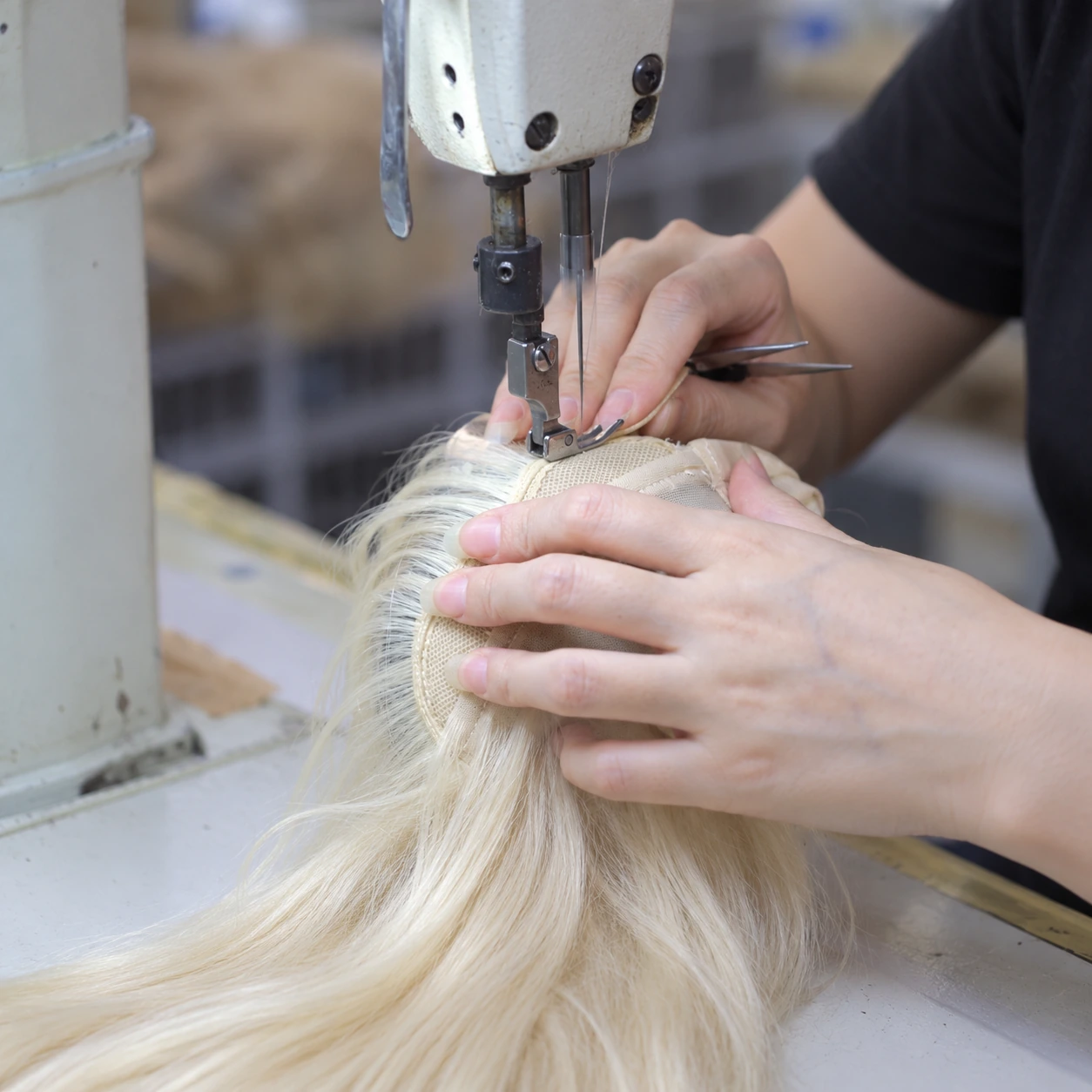 Hands guiding the beige mesh base of a light blonde hairpiece under an industrial sewing machine needle.