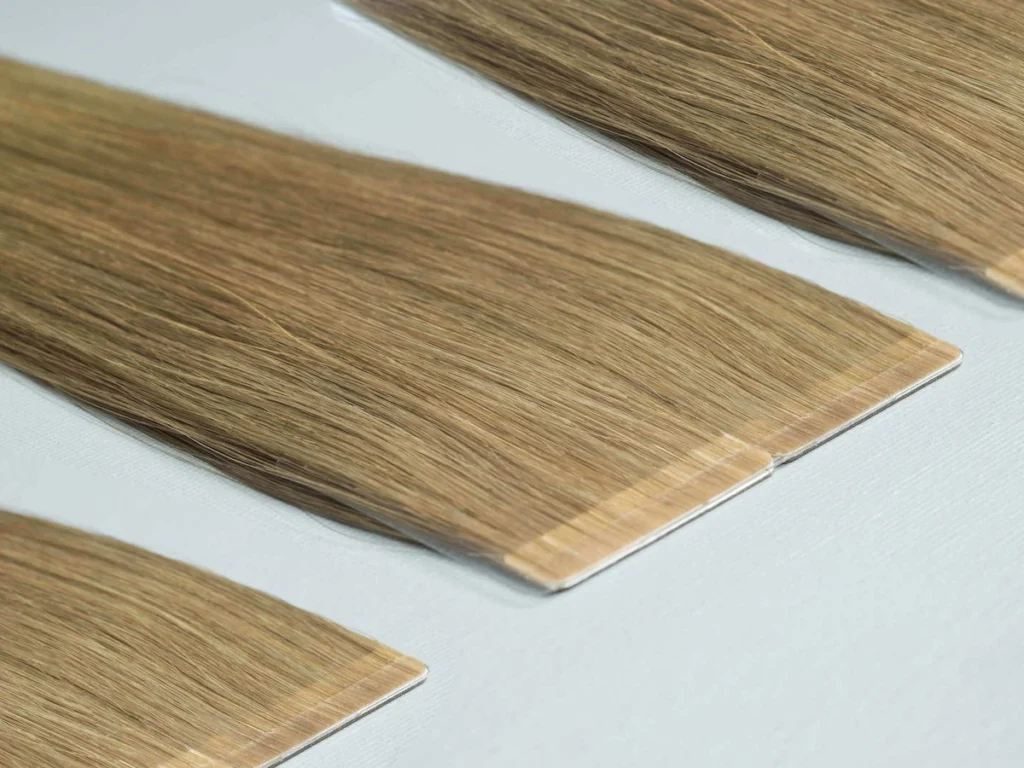 Slimline Tape-In hair extensions arranged diagonally on a pale background, showing narrow tape tabs and straight hair strands.