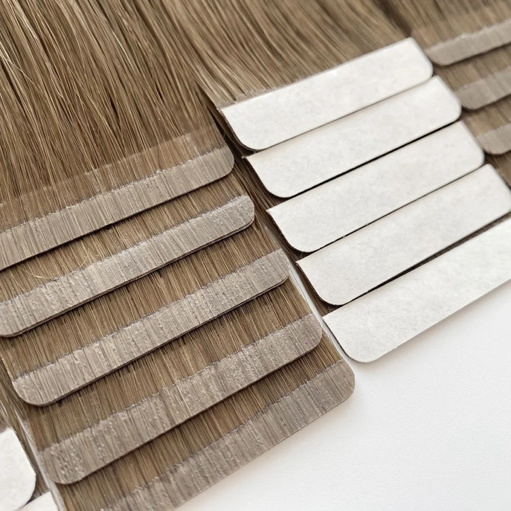 Detailed view of Slimline Tape-In hair extensions showing textured brown tape tabs beside stacked white release liners.