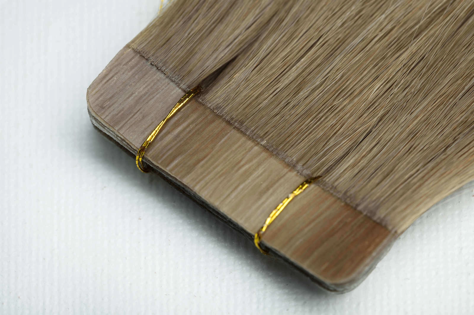 Two bundles of light brown tape-in hair extensions showing flat rectangular tabs secured with gold ties